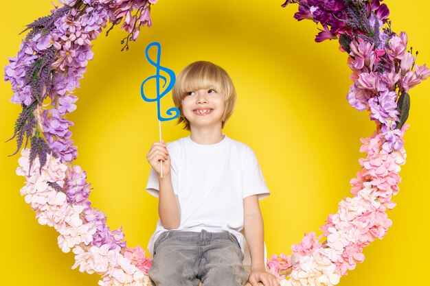 Ein blonder lächelnder Junge der Vorderansicht im weißen T-Shirt, das blaue Note hält, die auf der Blume sitzt, machte Stand auf dem gelben Raum