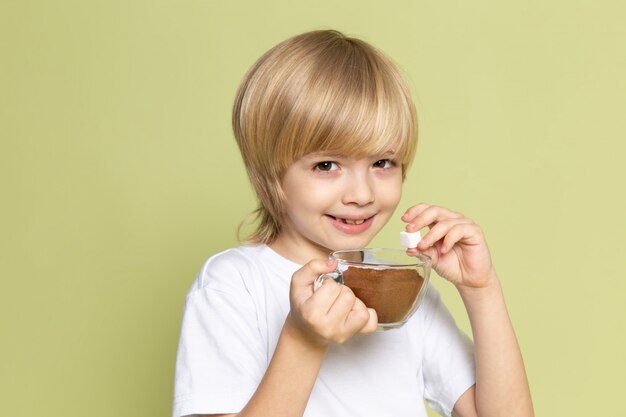 Ein blondes lächelndes Kind der Vorderansicht im weißen T-Shirt, das Kaffeepulver auf dem steinfarbenen Schreibtisch hält