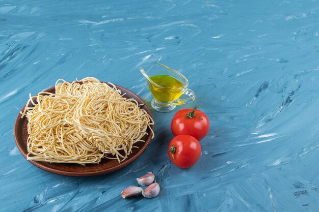 Ein brauner Teller mit rohen Nudeln mit zwei frischen roten Tomaten und Öl auf blauem Grund.