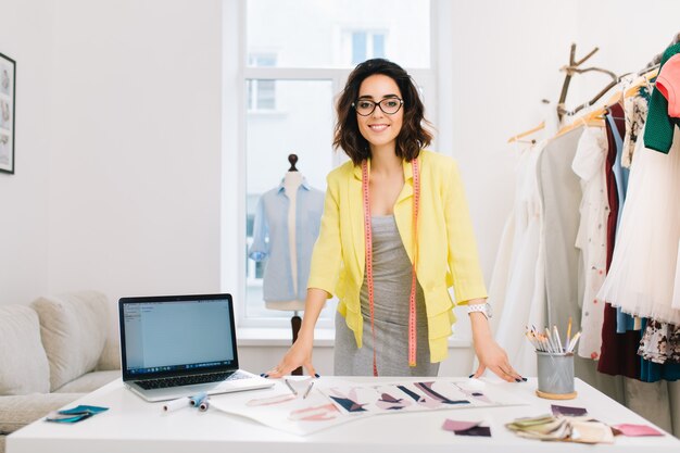 Ein brünettes Mädchen in einem grauen Kleid und einer gelben Jacke steht in einem Werkstattstudio neben dem Tisch. Sie hat viele kreative Sachen auf dem Tisch. Sie lächelt in die Kamera.