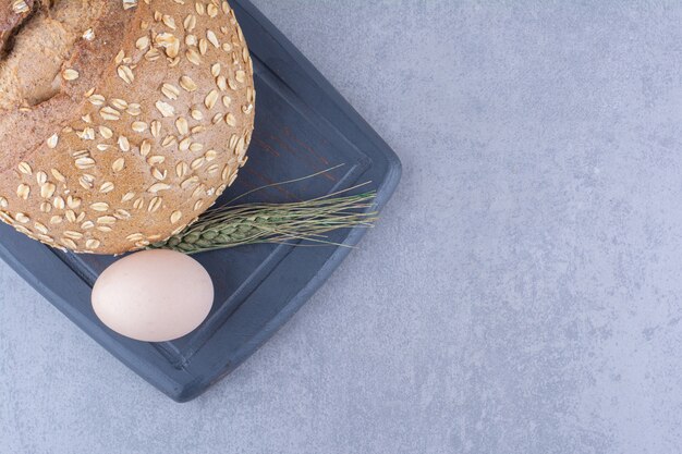 Ein Ei, ein Brot und ein einzelner Weizenhalm auf einem Brett auf Marmoroberfläche