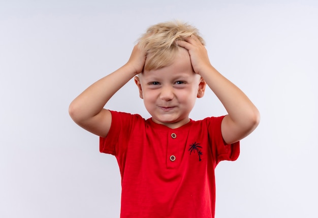 Ein entzückender kleiner blonder junge im roten t-shirt, das haar mit den fingern hält