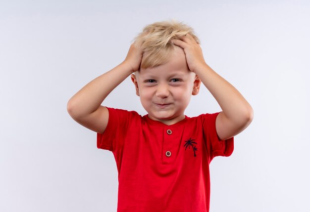 Ein entzückender kleiner blonder Junge im roten T-Shirt, das Haar mit den Fingern hält