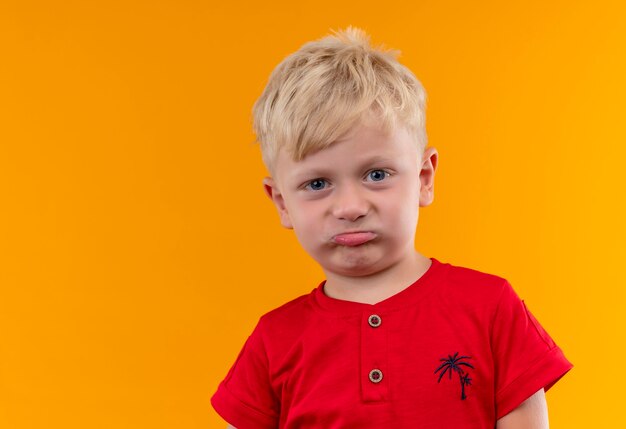 Ein entzückender kleiner Junge mit blonden Haaren und blauen Augen, die rotes T-Shirt tragen, das mit missfallenem Ausdruck schaut