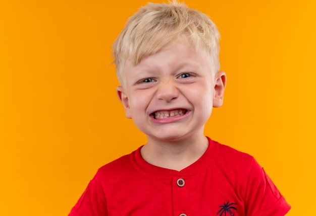 Ein entzückender kleiner Junge mit blonden Haaren und blauen Augen, die rotes T-Shirt tragen, das seine Zähne zeigt