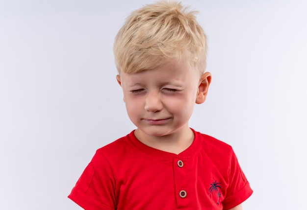 Ein entzückender kleiner süßer Junge mit blondem Haar, das rotes T-Shirt trägt, das seine Augen schließt