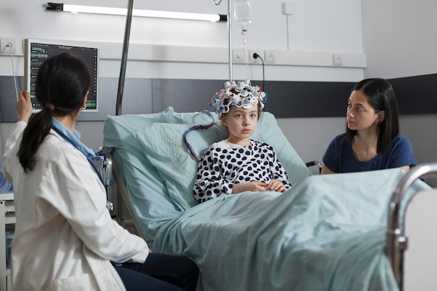 Ein erfahrener Kinderarzt überwacht den Zustand des Gehirns eines Mädchens im Krankenhaus mit einem EEG-Headset. Neurologiespezialist, der die neurologische Krankheitsentwicklung eines kranken Kindes misst, das in einer pädiatrischen Gesundheitseinrichtung sitzt.