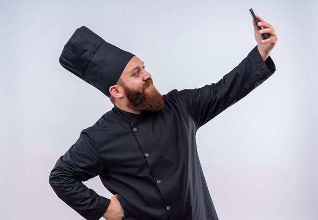 Ein erfreuter bärtiger Kochmann in der schwarzen Uniform, die selfie mit Handy auf einer weißen Wand nimmt