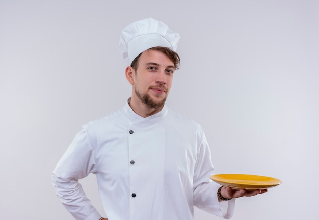 Ein erfreuter junger bärtiger Kochmann in der weißen Uniform, die einen gelben Teller bereit zum Essen darstellt, während auf einer weißen Wand betrachtet