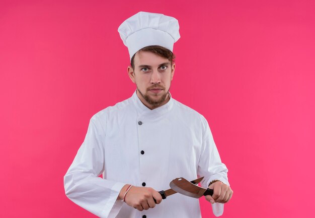 Ein ernsthafter junger bärtiger Kochmann in der weißen Uniform, die das Messer des Kochmützenschärfens beim Betrachten auf einer rosa Wand trägt