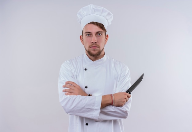 Ein ernsthafter junger bärtiger Kochmann in der weißen Uniform, die ein Messer hält, während auf einer weißen Wand schaut