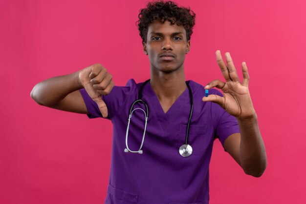 Ein ernsthafter junger gutaussehender dunkelhäutiger Arzt mit lockigem Haar in violetter Uniform mit Stethoskop, das beim Halten der Pille die Daumen nach unten zeigt