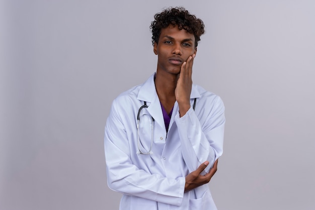 Ein ernsthafter junger hübscher dunkelhäutiger männlicher Arzt mit lockigem Haar, der weißen Kittel mit Stethoskop trägt, das Hand auf Wange hält