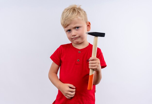 Ein ernsthafter kleiner süßer blonder Junge im roten T-Shirt, der Hammer hält, während auf einer weißen Wand schaut