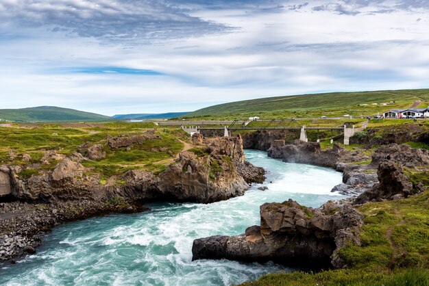 Ein Fluss von Godafoss Falls, Akureyri, Island, umgeben von riesigen Felsen und einer Betonbrücke