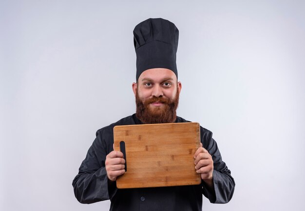 Ein freudiger bärtiger Kochmann in der schwarzen Uniform, die hölzerne Küchenbrett zeigt, während auf einer weißen Wand schaut