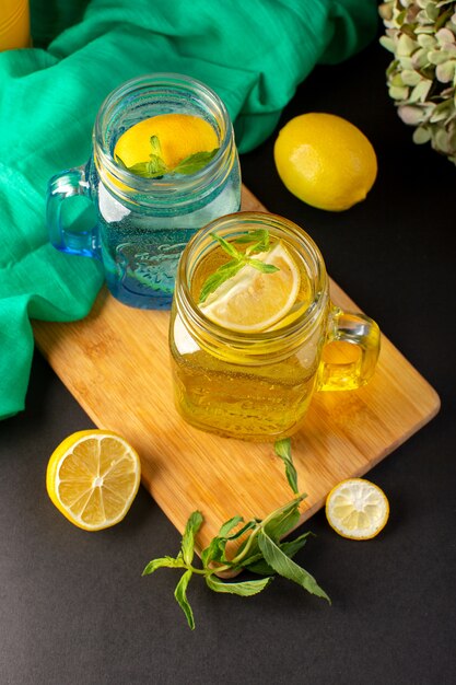 Ein frisches kühles Getränk des Zitronencocktails der Vorderansicht in geschnittenen Glasbechern und ganzen Zitronen zusammen mit Blumen auf der dunklen Hintergrundcocktailgetränkfrucht