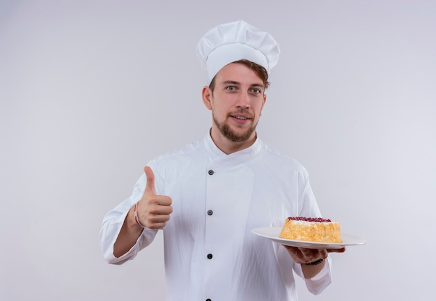 Ein fröhlicher junger bärtiger kochmann, der weiße kochuniform und hut hält, einen teller mit kuchen hält und daumen oben zeigt, während er auf eine weiße wand schaut