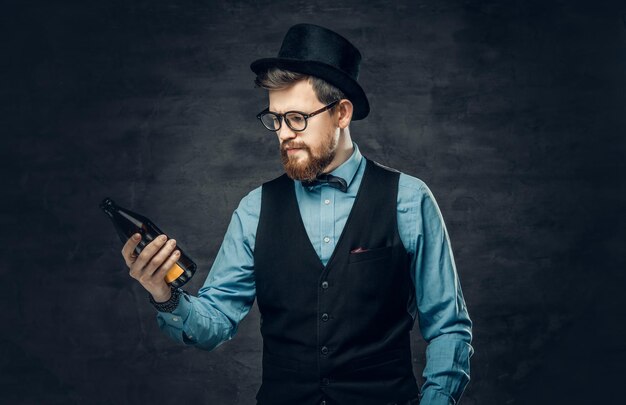 Ein funky bärtiger Hipster-Mann in einem blauen Hemd, einer eleganten Weste und einem Zylinder hält eine Craft-Bierflasche.