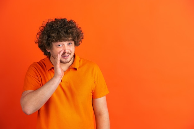 Ein Geheimnis flüstern. Das monochrome Portrait des kaukasischen jungen Mannes auf oranger Wand. Schönes männliches lockiges Modell im lässigen Stil.