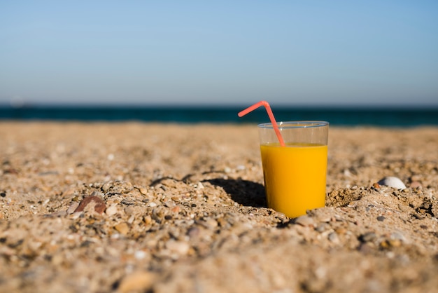 Ein Glas gelber Saft mit rotem Trinkhalm im Sand am Strand