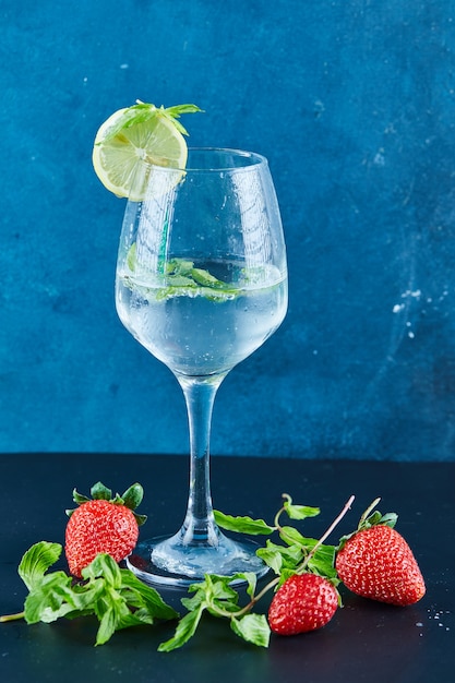 Ein Glas Saft mit Minze und Zitronenscheibe auf blauer Oberfläche mit frischen Erdbeeren und Minze