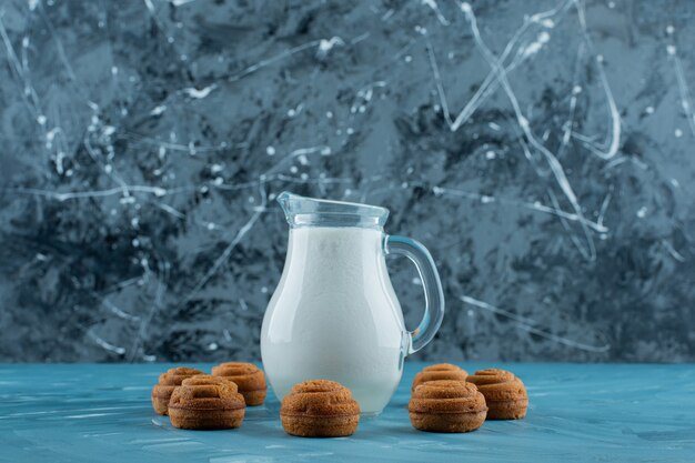 Ein Glaskrug frische Milch mit süßen runden Keksen auf einer blauen Oberfläche