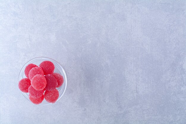 Ein Glasteller voller roter zuckerhaltiger Fruchtgummibonbons auf grauer Oberfläche