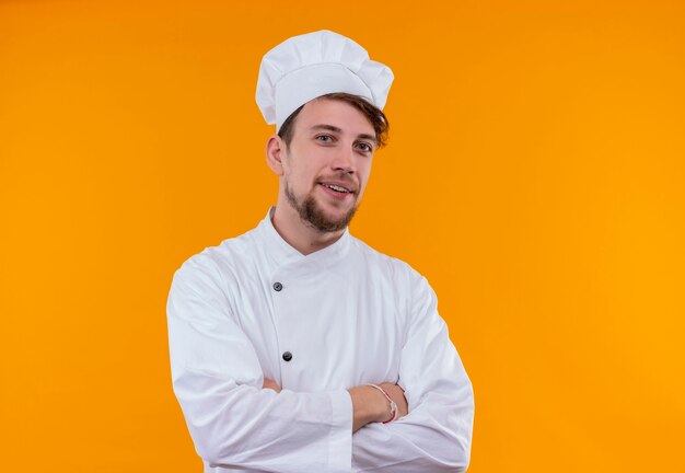 Ein glücklicher hübscher junger bärtiger Kochmann in der weißen Uniform, die Händchen haltend hält, während auf einer orange Wand schaut