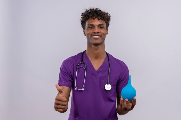 Ein glücklicher junger gutaussehender dunkelhäutiger Arzt mit lockigem Haar in violetter Uniform mit Stethoskop, das Daumen hoch zeigt, während er einen Einlauf hält