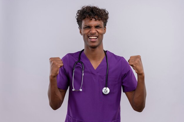 Ein glücklicher junger gutaussehender dunkelhäutiger Arzt mit lockigem Haar in violetter Uniform mit Stethoskop und geballter Faust