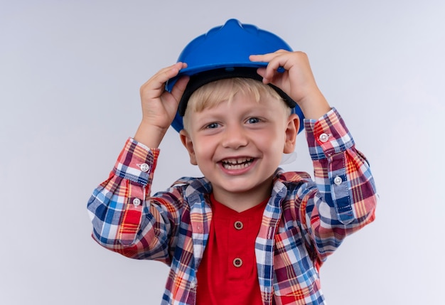 Ein glücklicher kleiner Junge mit blondem Haar trägt kariertes Hemd, das seinen blauen Helm hält, während er auf eine weiße Wand schaut