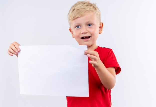 Ein glücklicher kleiner süßer blonder Junge im roten T-Shirt lächelnd und zeigt leeres Blatt Papier auf einer weißen Wand