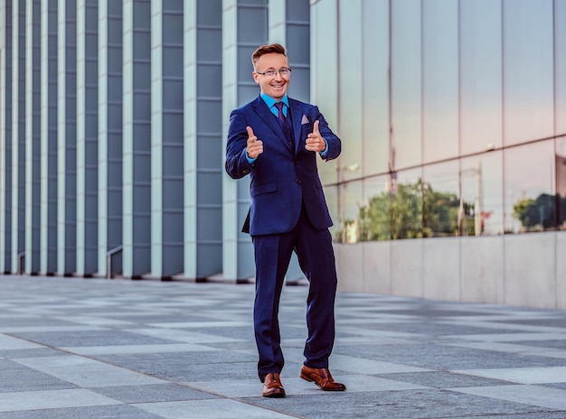 Ein glücklicher, selbstbewusster Geschäftsmann in einem eleganten Anzug zeigt eine coole Geste, während er im Freien vor dem Hintergrund der Stadt steht.