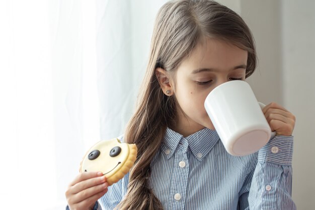 Ein Grundschulmädchen frühstückt mit Milch und lustigen Keksen in Form eines Smileys.