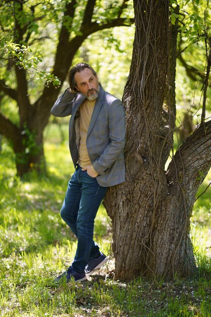 Ein gut aussehender, reifer Vogelmann in lässigem Stehen unter dem Baum auf dem Gras, der mit den Händen in den Taschen hinter den Baum schaut Freiberufler verbringen Zeit mit der Arbeit im Freien