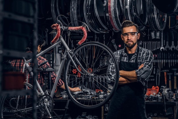 Ein gutaussehender bärtiger Mann mit Brille steht in seiner eigenen Werkstatt in der Nähe eines festen Fahrrads.