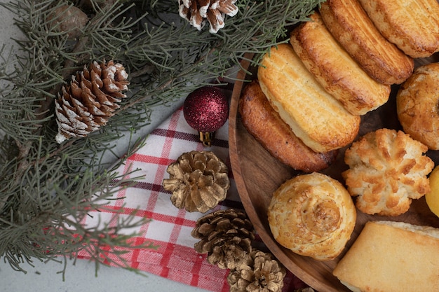 Ein Holzteller voller süßem Gebäck mit Weihnachtskugeln und Tannenzapfen