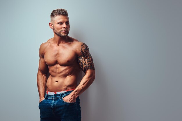 Ein hübscher, hemdloser Bodybuilder mit stylischem Haarschnitt und Bart, mit Tätowierung auf dem Arm, der in einem Studio posiert. Isoliert auf grauem Hintergrund.