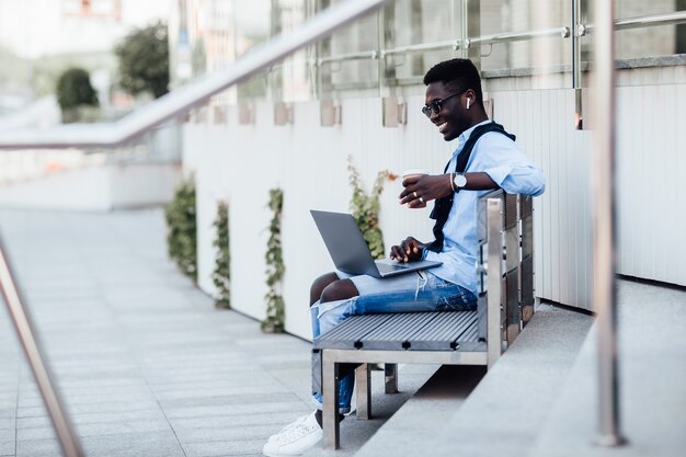 Ein hübscher junger Geschäftsmann, der mit seinem Laptop auf einer sonnigen Straße neben einem Park auf einer Bank sitzt. Mit Tasse Kaffee.
