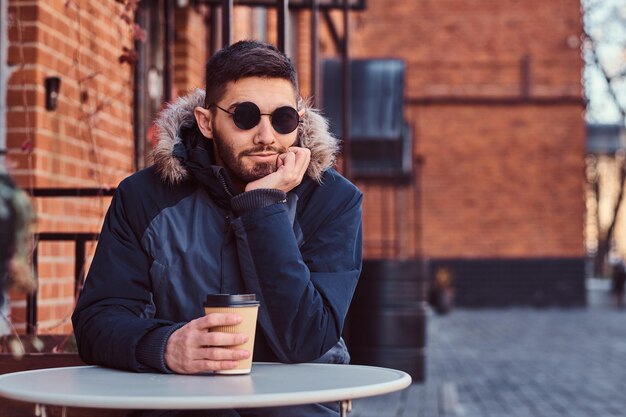 Ein hübscher junger Mann, der einen Wintermantel trägt, sitzt mit Kaffee im Café im Freien.