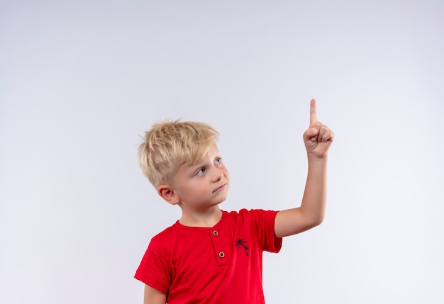 Ein hübscher kleiner Junge mit blonden Haaren und blauen Augen, die rotes T-Shirt tragen, das mit Zeigefinger auf einer weißen Wand zeigt