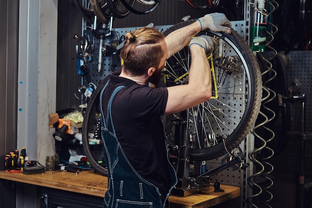 Ein hübscher rothaariger Mann in einem Jeansoverall, der in einer Reparaturwerkstatt mit einem Fahrradrad arbeitet. Ein Arbeiter entfernt den Fahrradreifen in einer Werkstatt.