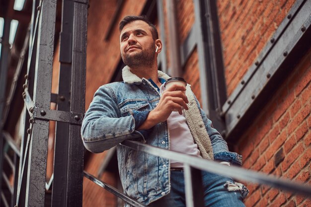 Ein hübscher, stilvoller Mann, der eine Jeansjacke auf der Treppe draußen trägt und einen Kaffee zum Mitnehmen hält.