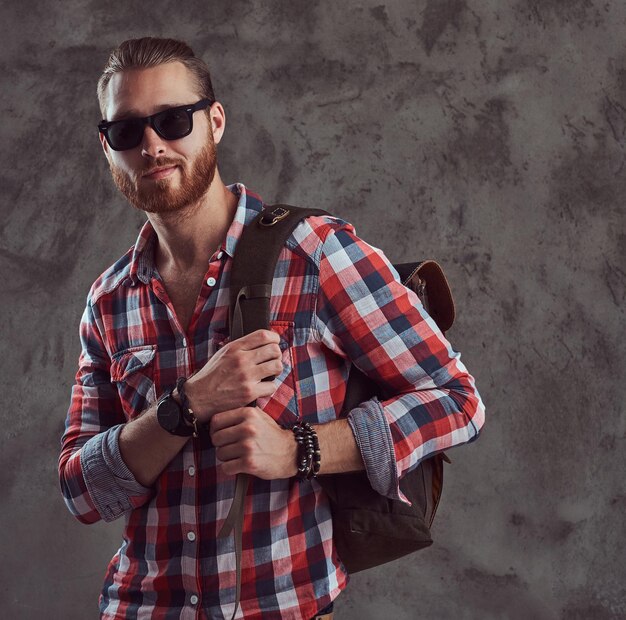 Ein hübscher, stylischer rothaariger Reisender in Flanellhemd und Sonnenbrille mit Rucksack, der in einem Studio auf grauem Hintergrund posiert.