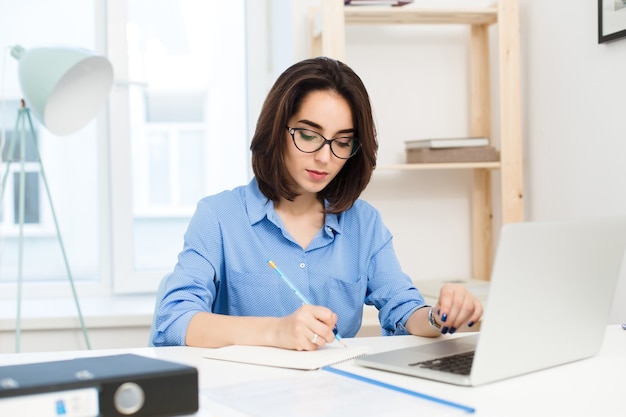 Ein hübsches brünettes Mädchen arbeitet am Tisch im Büro. Sie trägt ein blaues Hemd und eine schwarze Brille. Sie schreibt ernst.