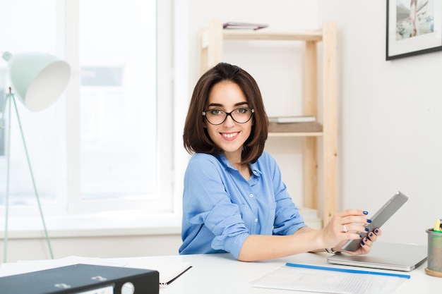 Ein hübsches brünettes Mädchen im blauen Hemd sitzt am Tisch im Büro. Sie arbeitet mit einem Laptop und lächelt in die Kamera.