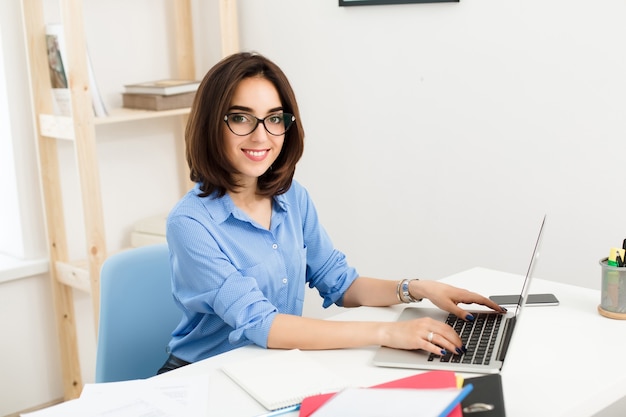Ein hübsches brünettes Mädchen sitzt und tippt auf einem Laptop am Tisch im Büro. Sie lächelt freundlich in die Kamera. Sie trägt ein blaues Hemd und eine schwarze Brille.