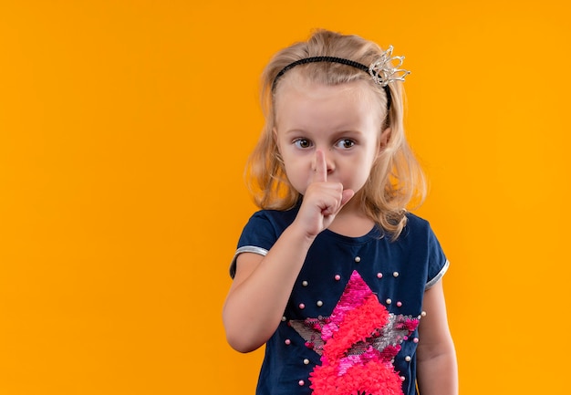 Ein hübsches kleines Mädchen, das ein dunkelblaues Hemd im Kronenstirnband trägt, das shh Geste mit Zeigefinger auf Mund zeigt und Seite auf einer orange Wand schaut