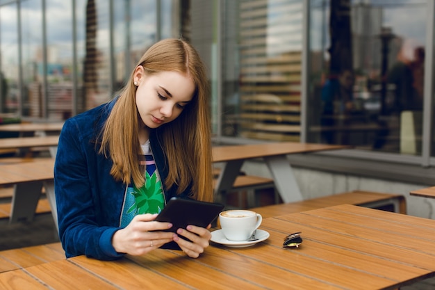 Ein hübsches Mädchen mit langen Haaren sitzt am Tisch auf der Terrasse in der Stadt. Sie arbeitet an dem Tablet.